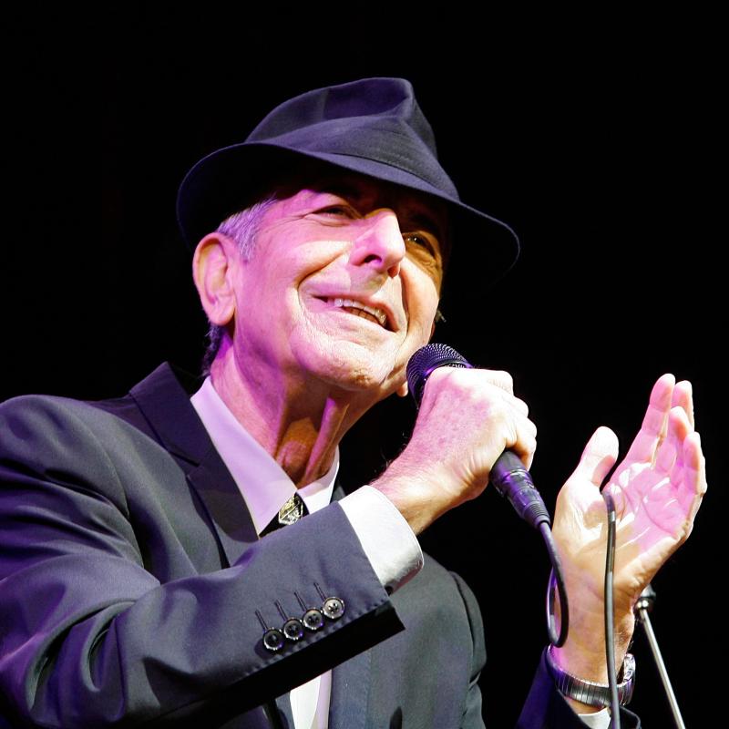 Musician Leonard Cohen singing on stage in a black suit and hat