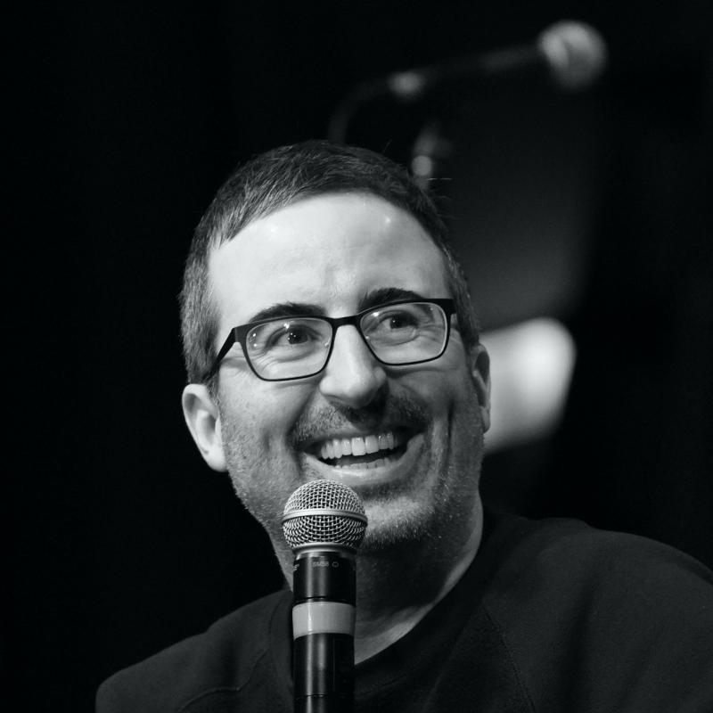Comedian John Oliver laughs in front of a microphone in black and white.