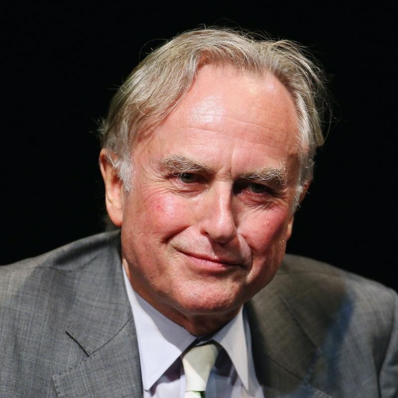 Professor Richard Dawkins smirks on stage and looks off-camera while wearing a suit
