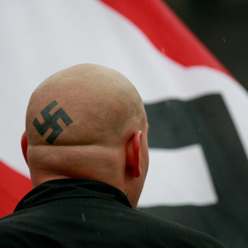 A white American skinhead with a swastika tattoo on the back of his head faces away from the camera and looks at a Nazi flag
