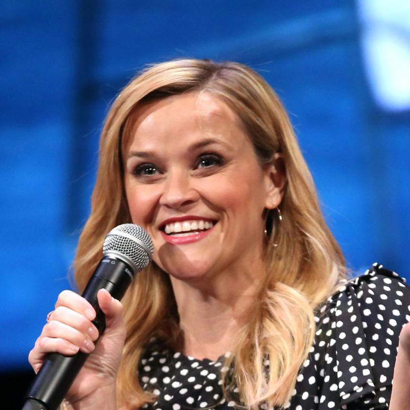 Actor Reese Witherspoon speaks into a mic while smiling on stage