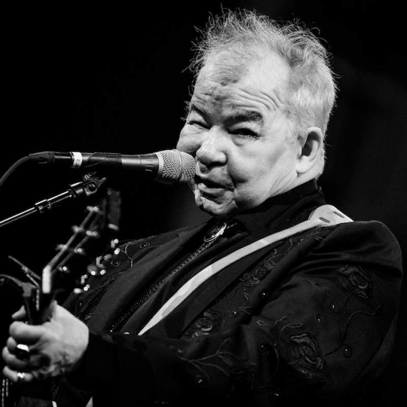 John Prine plays his guitar and looks at the camera during a live performance