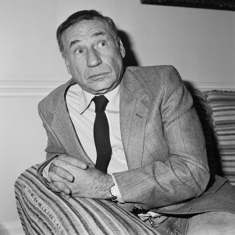 Filmmaker Mel Brooks reclines on a couch in a suit in this black and white image from 1984