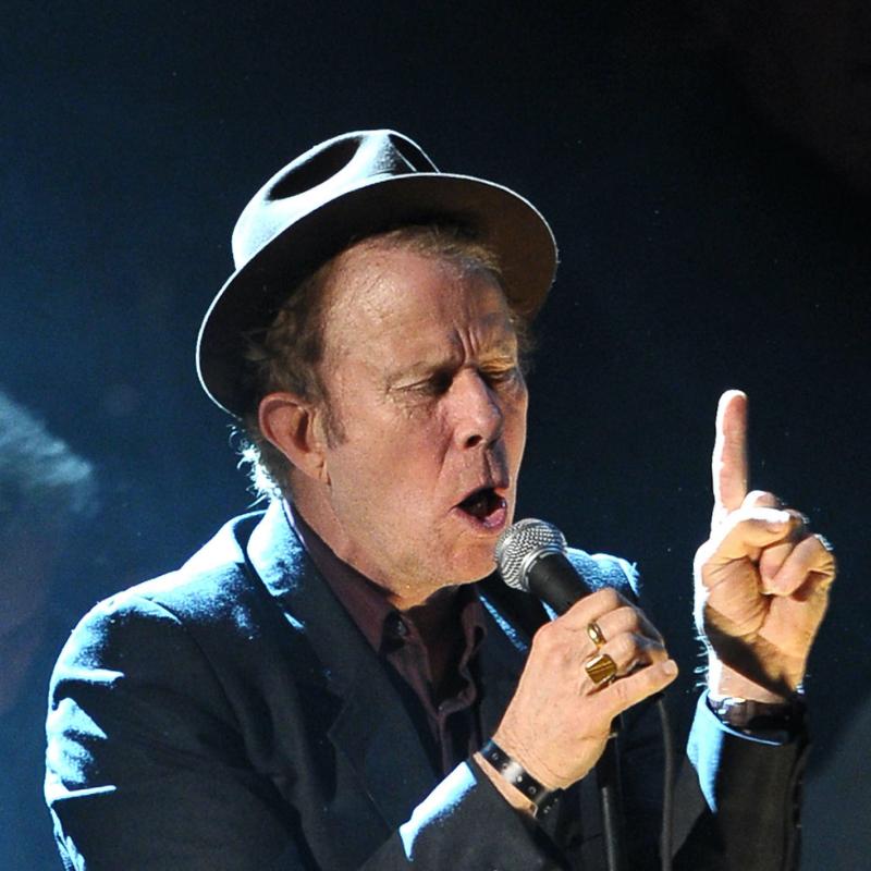 Singer and musician Tom Waits sings into a mic on stage with a hat tipped back on his head