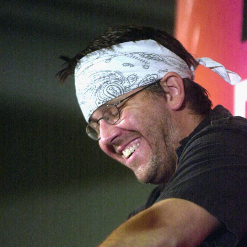 Author David Foster Wallace smiles while speaking at a lectern wearing a white bandana