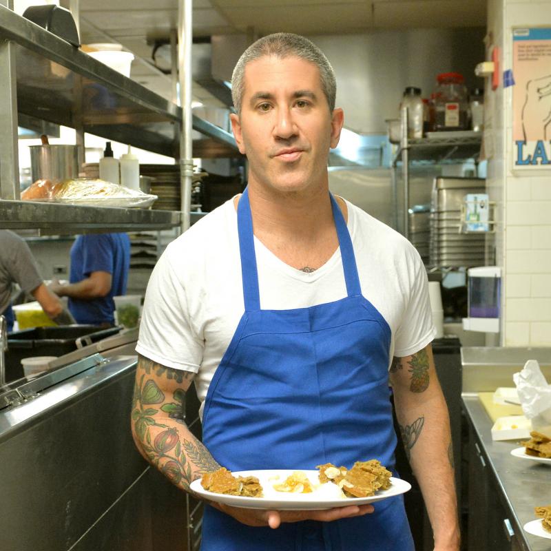 Chef Michael Solomonov holds a plate of food in his kitchen while dressed in a blue apron